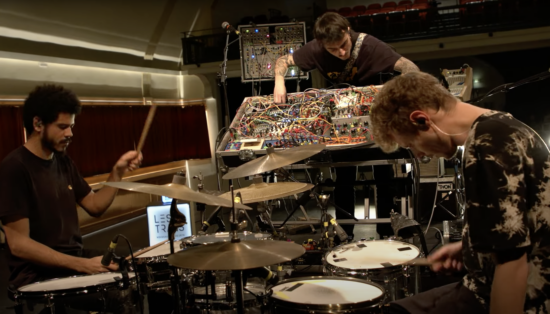 Deux artistes jouent de la batterie face à face, un troisième manipule les boutons et les câbles d'une grande console sonore. Ils sont en studio.