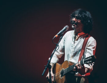 Un chanteur-guitariste chevelu et moustachu sur scène en train de jouer et de fumer une cigarette