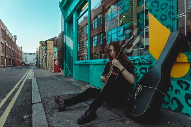 Dans la rue, une jeune femme guitariste joue assise au sol appuyée contre une vitrine colorée