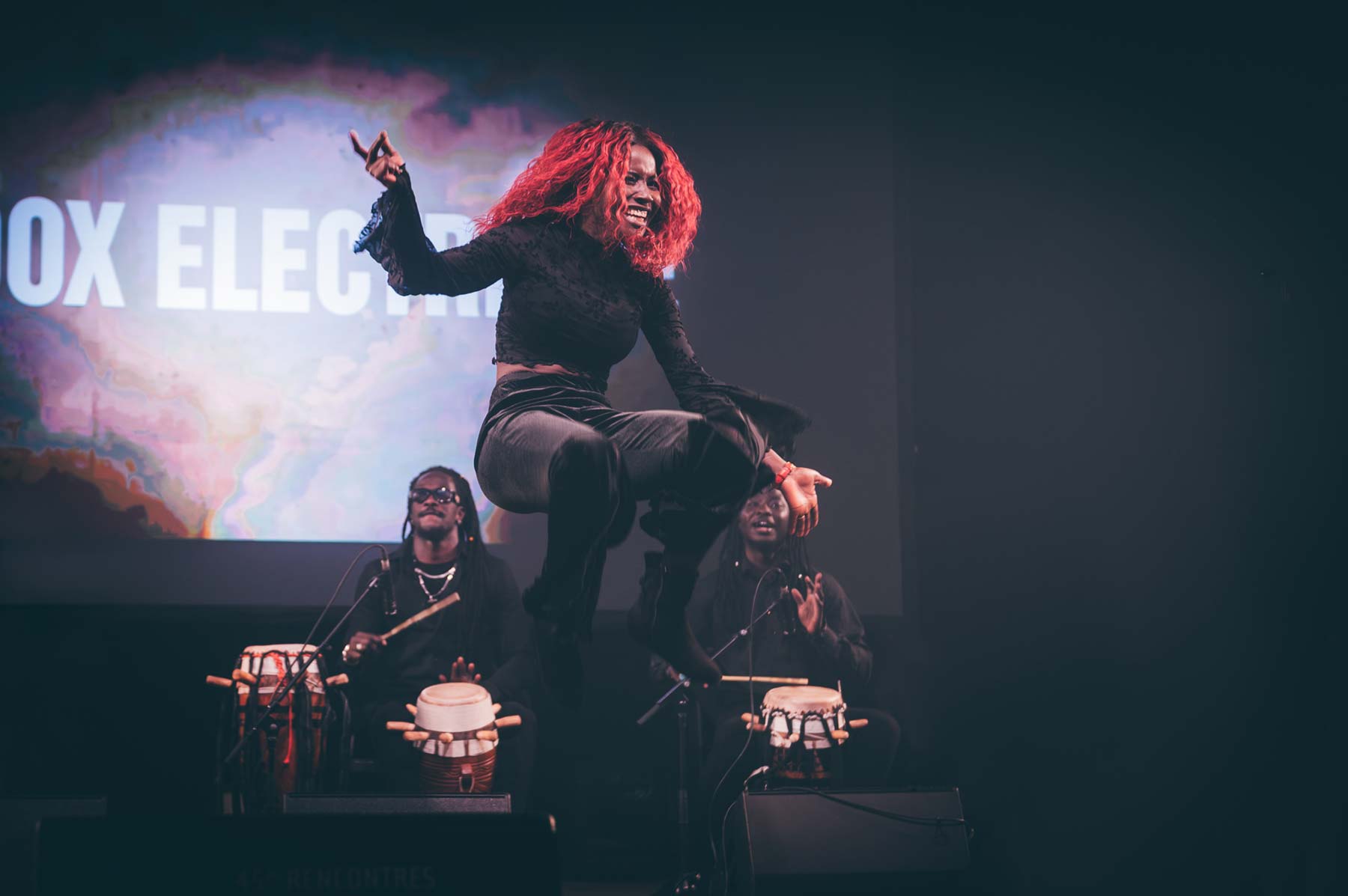 une artiste au cheveux rouge en plein saut devant des joueurs de percussion