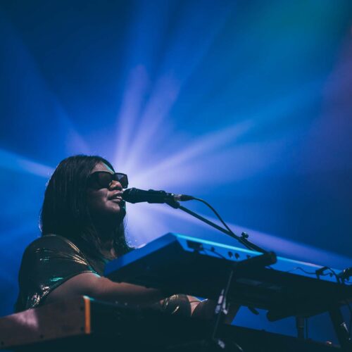 Une artiste avec des lunettes noire souriante joue du clavier en chantant